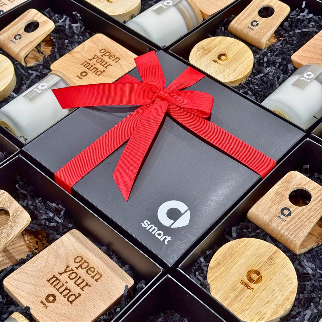 A corporate gift box arrangement featuring a central black box tied with a bright red ribbon and printed with the 'smart' logo, surrounded by multiple open black boxes containing small wooden electronic devices and round wooden lids on a black shredded paper background.