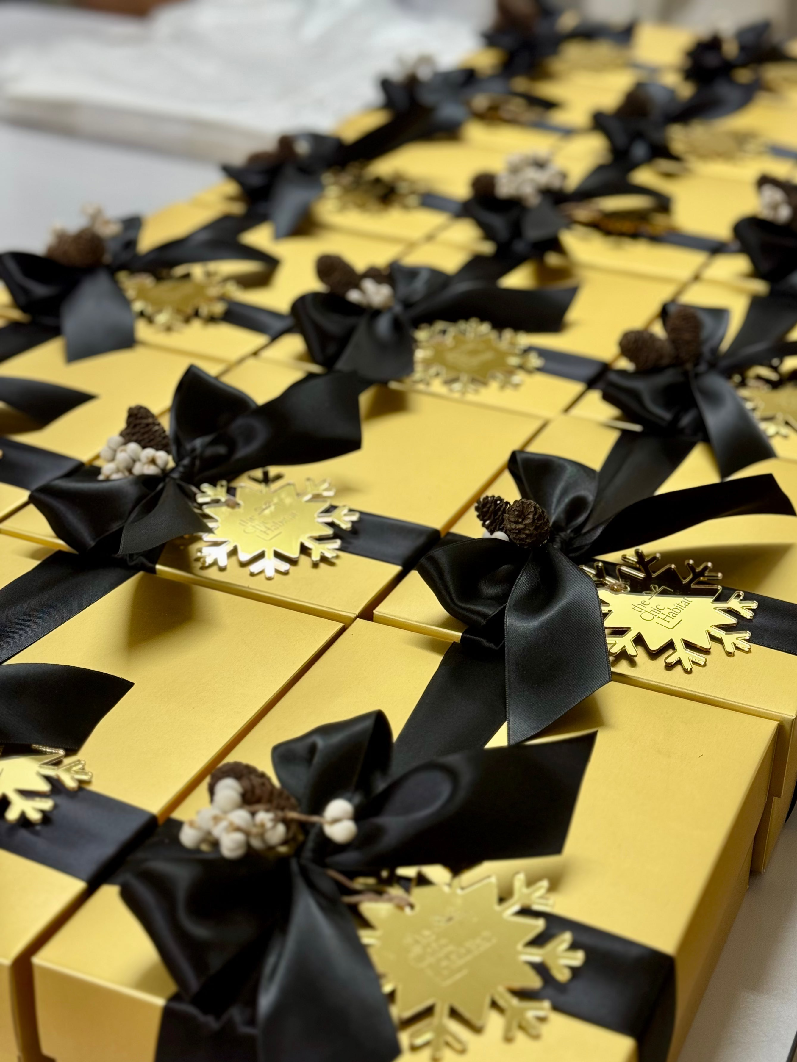 A close-up view of multiple gold Christmas gift boxes arranged on a table, each tied with a black satin ribbon and decorated with a gold snowflake and small dried floral accents.