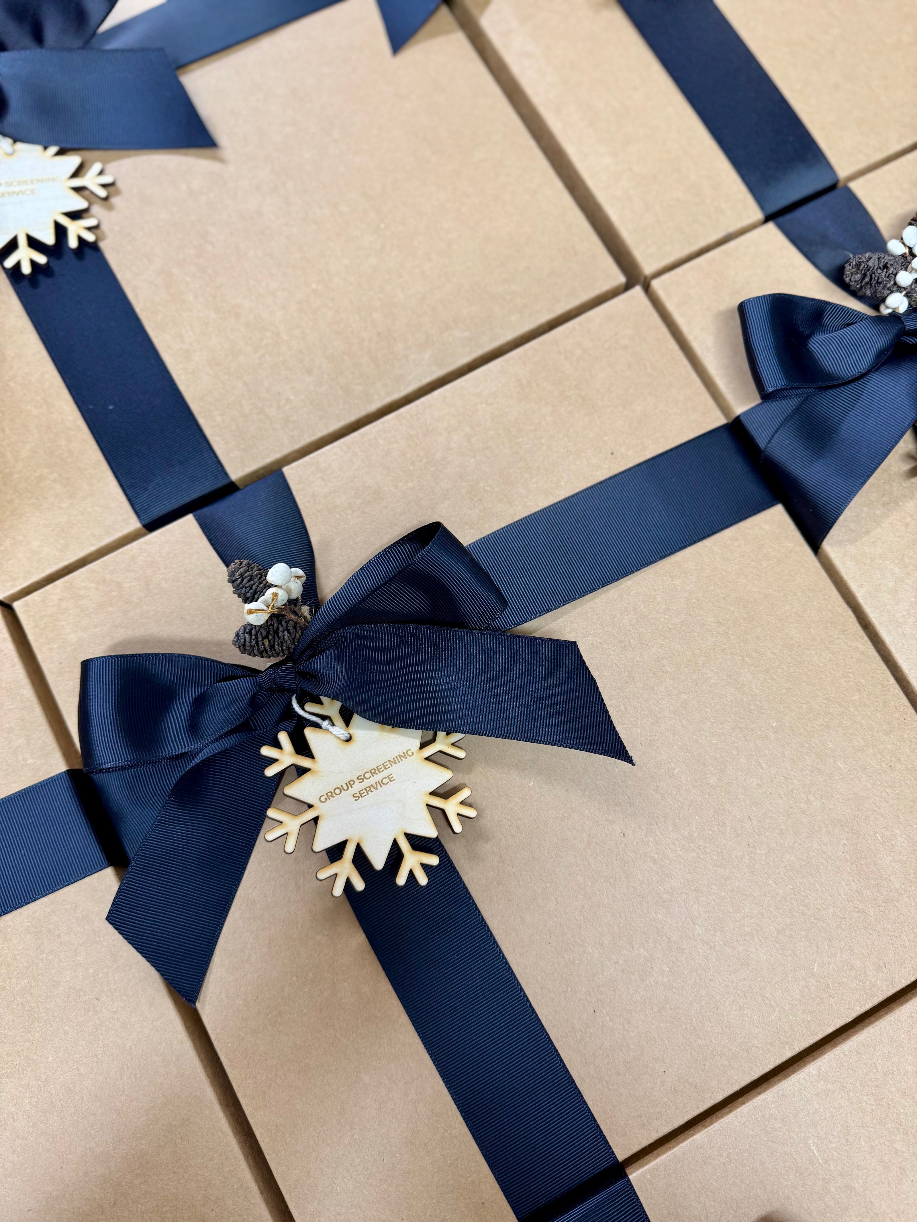 A close-up of several kraft paper gift boxes tied with navy blue satin ribbons and bow, each decorated with a white snowflake ornament and small pinecones.