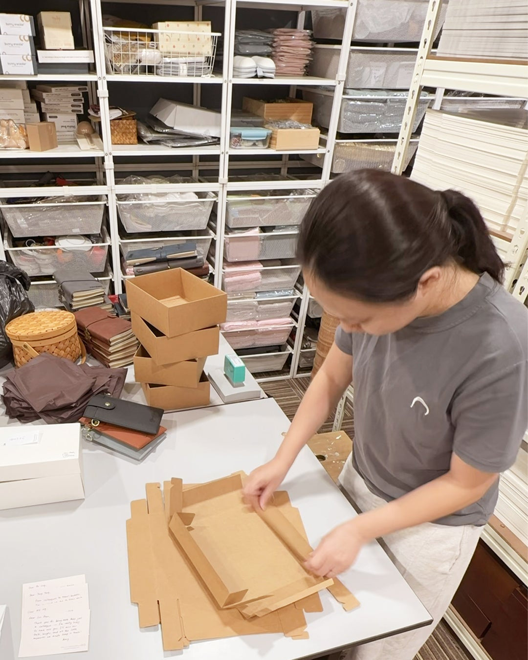 A woman in a gray t-shirt and white pants is shown packing or organizing brown cardboard boxes on a white table inside a storage room with shelves of fabric or material samples in the background.